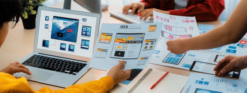 Team reviewing website wireframes and front-end web design layouts on laptops and printed mockups during a website planning session.